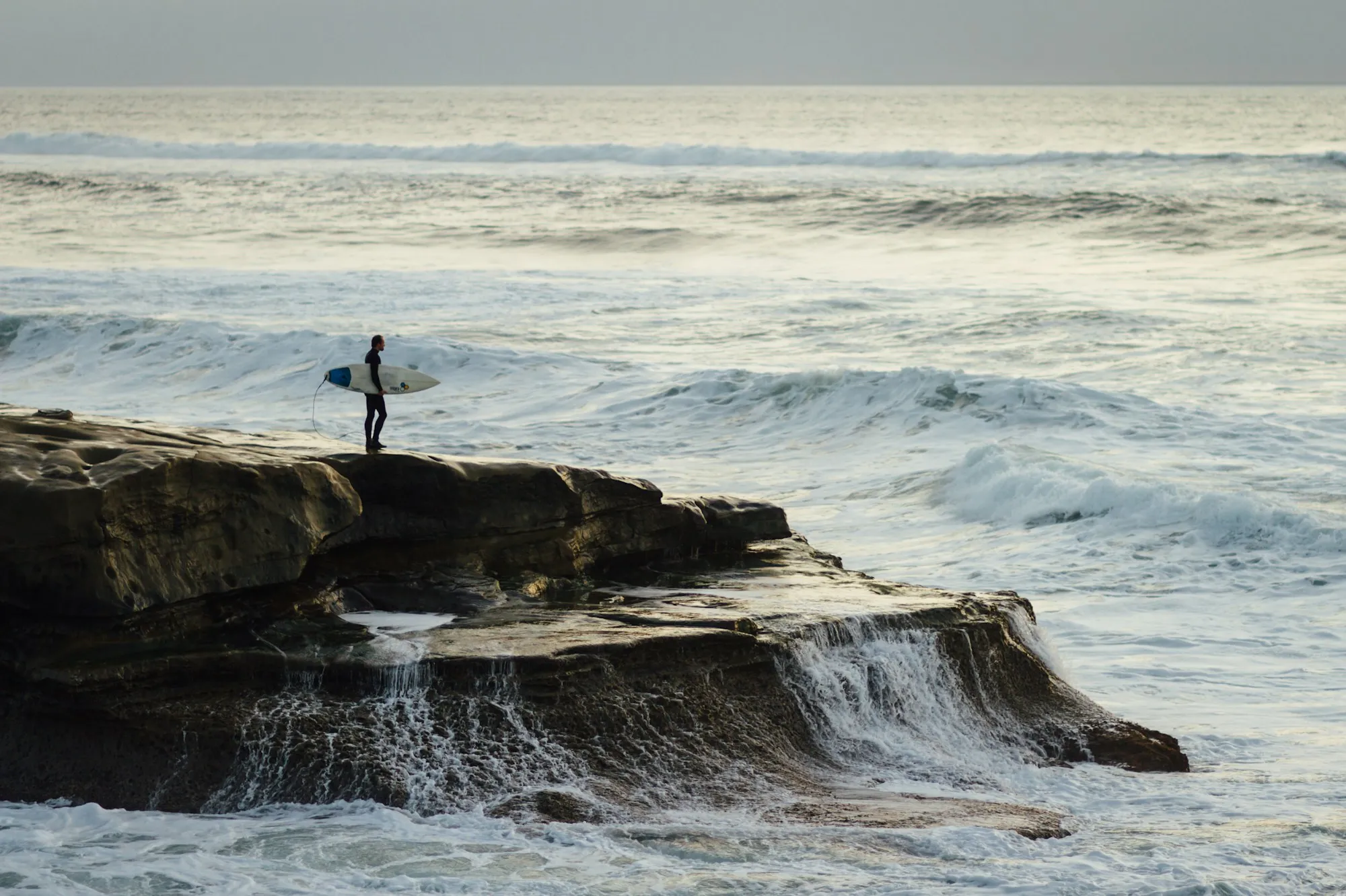A surfer carrying a board across a rock ledge beside breaking waves